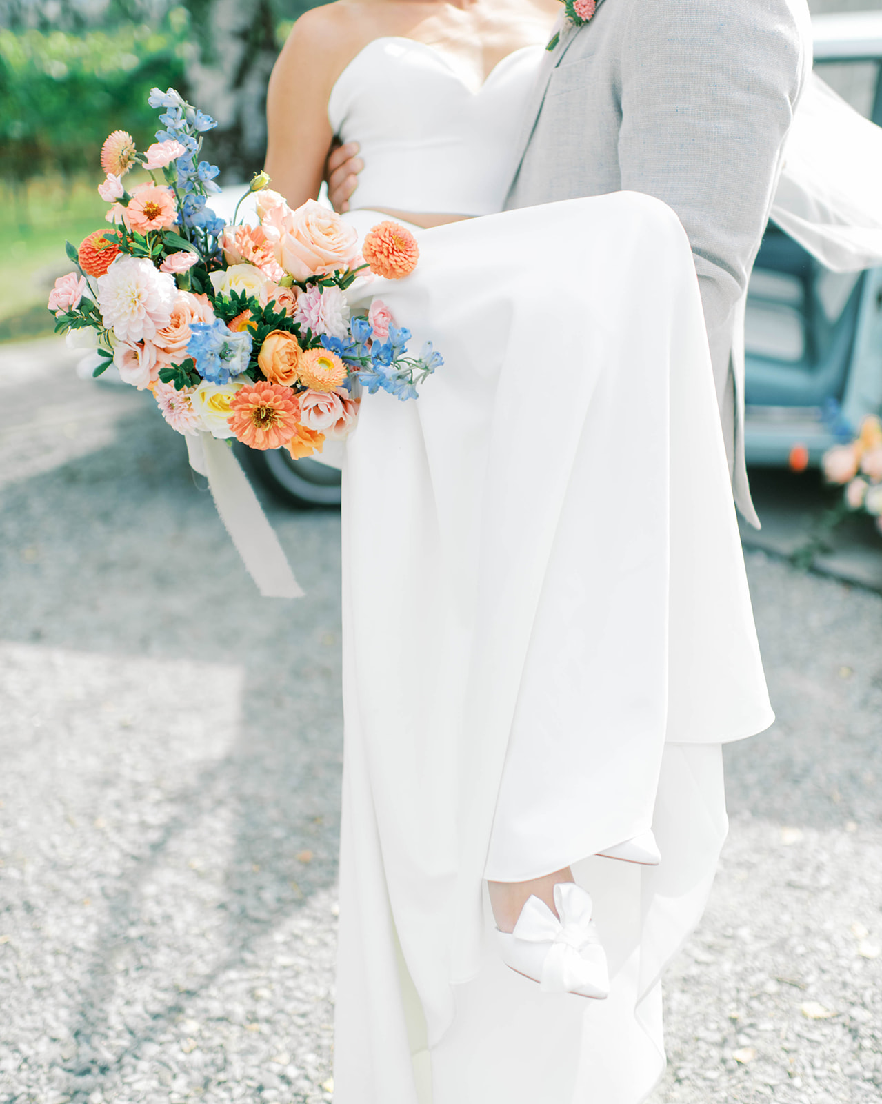 Bright summer florals in bridal bouquet adds a pop of colour to an enchanting Okanagan wedding, a glimpse of a vintage baby blue getaway car, groom carries bride in romantic couple's portrait 