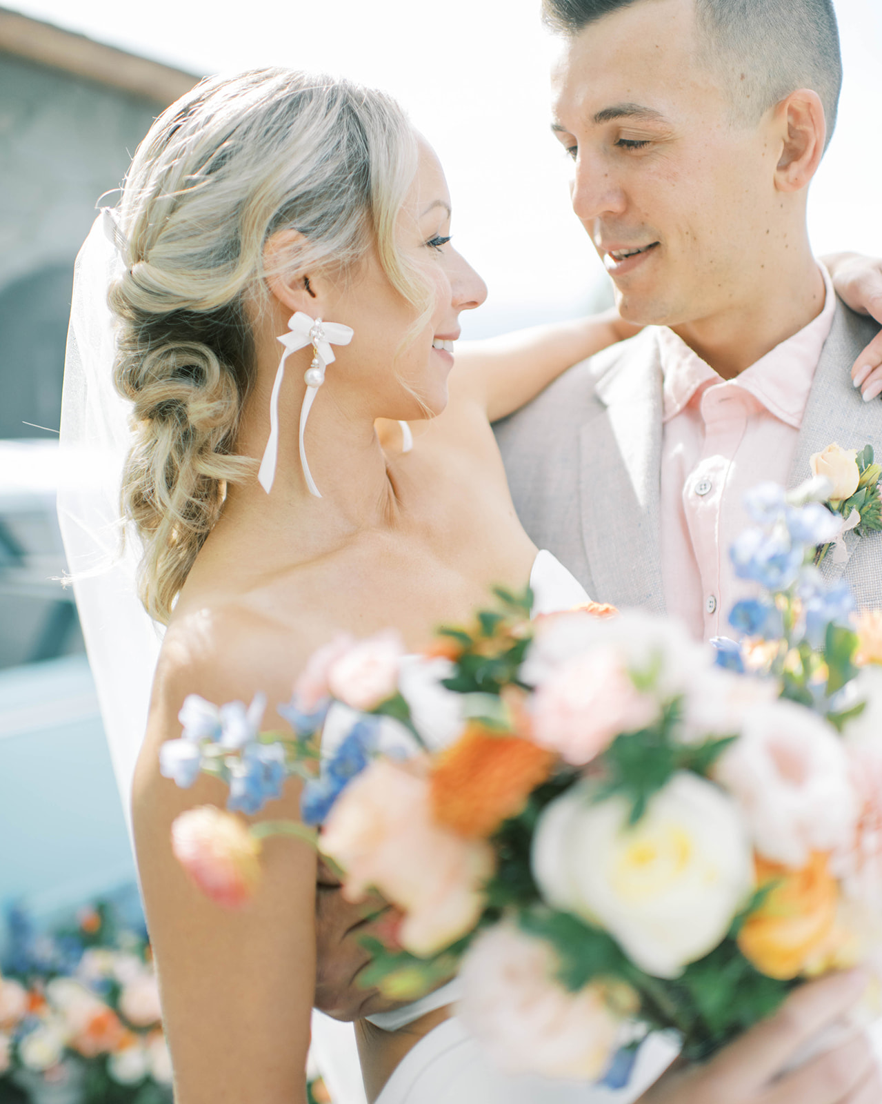 Loose pulled back bridal hairstyle showing off white bow earrings by Lindsay Marie Design in couple's portrait, pink shirt and bow tie worn by the groom to compliment the summer season, lush summery floral inspiration
