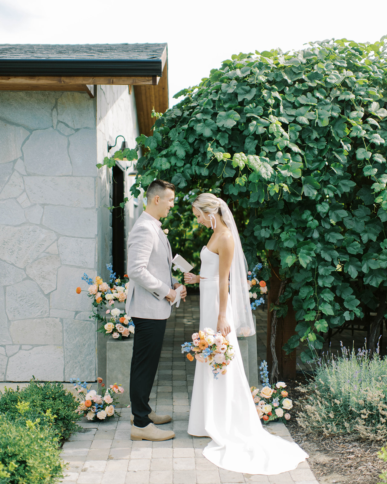 Stunning pedestal arch arrangement adorned with orange and blue summer wedding florals, creating an elegant backdrop for ceremony, Okanagan wedding at a Priest Creek Winery, couple dressed in modern and elegant wedding attire reading their vows to each other 