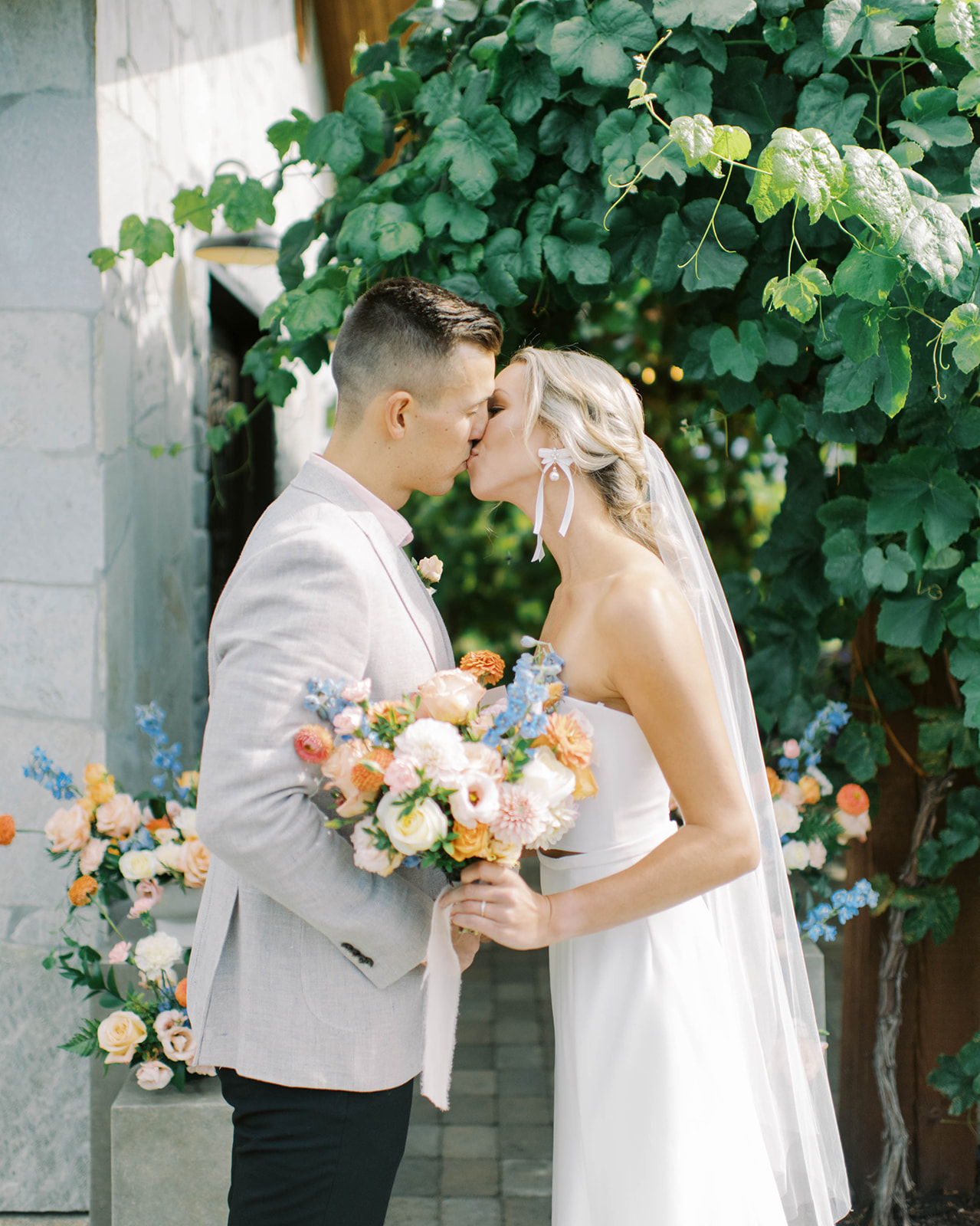 bride and groom share first kiss after reading their vows during a secluded ceremony at a Kelowna winery, bride is wearing a modern simple two-piece strapless wedding dress by Lillian Wild Bridal, bridal bouquet features summer florals in a pink, orange and blue colour palette