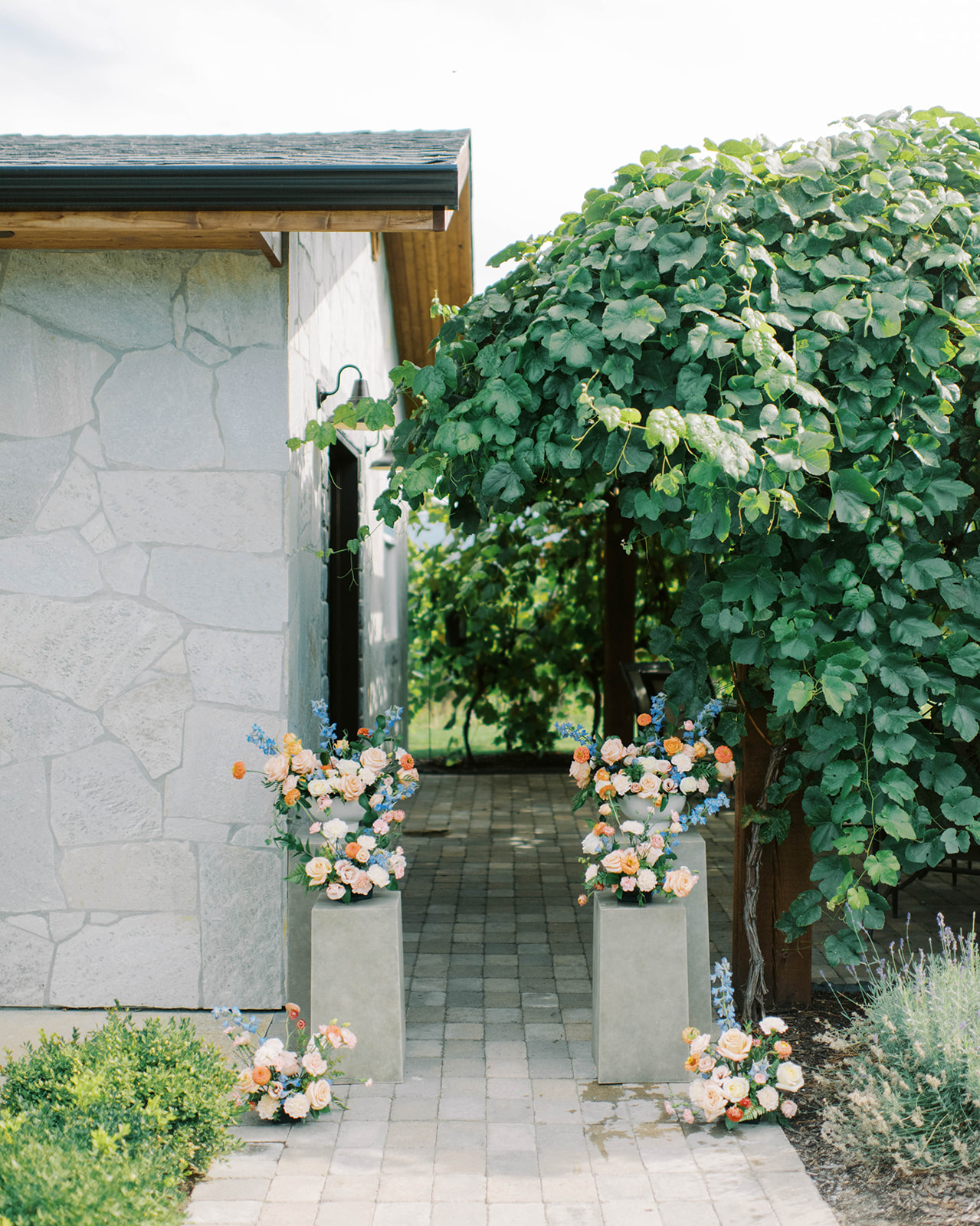 Pedestal stands adorned with beautiful floral arrangements, perfect for a summer wedding ceremony, outdoor ceremony with stone path aisle and lush green canopy at Kelowna winery