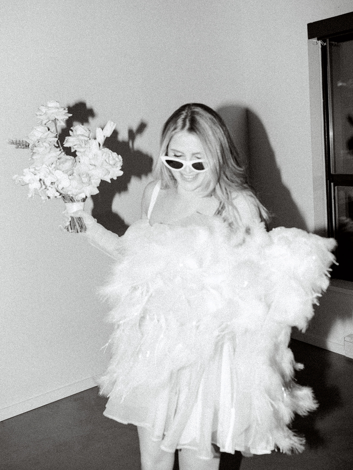 Black and white bridal portrait giving off a vintage look, cheeky bride holding bouquet wearing a white feather mini dress and white retro sunglasses for this modern photoshoot, black and white photography ideas for wedding