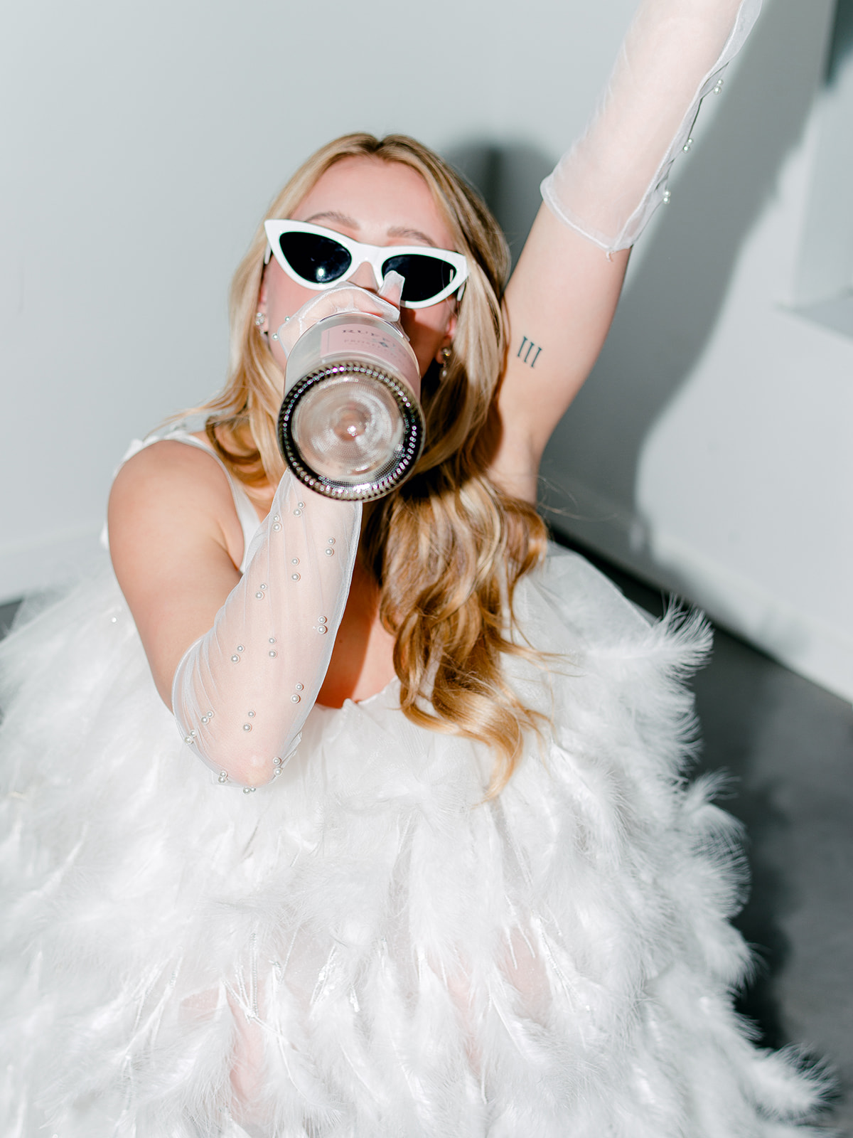 Modern and playful bride drinking champagne from the bottle in bridal portraits, sheer tulle bridal gloves with pearl details and white retro sunglasses worn by chic bride in flash photoshoot