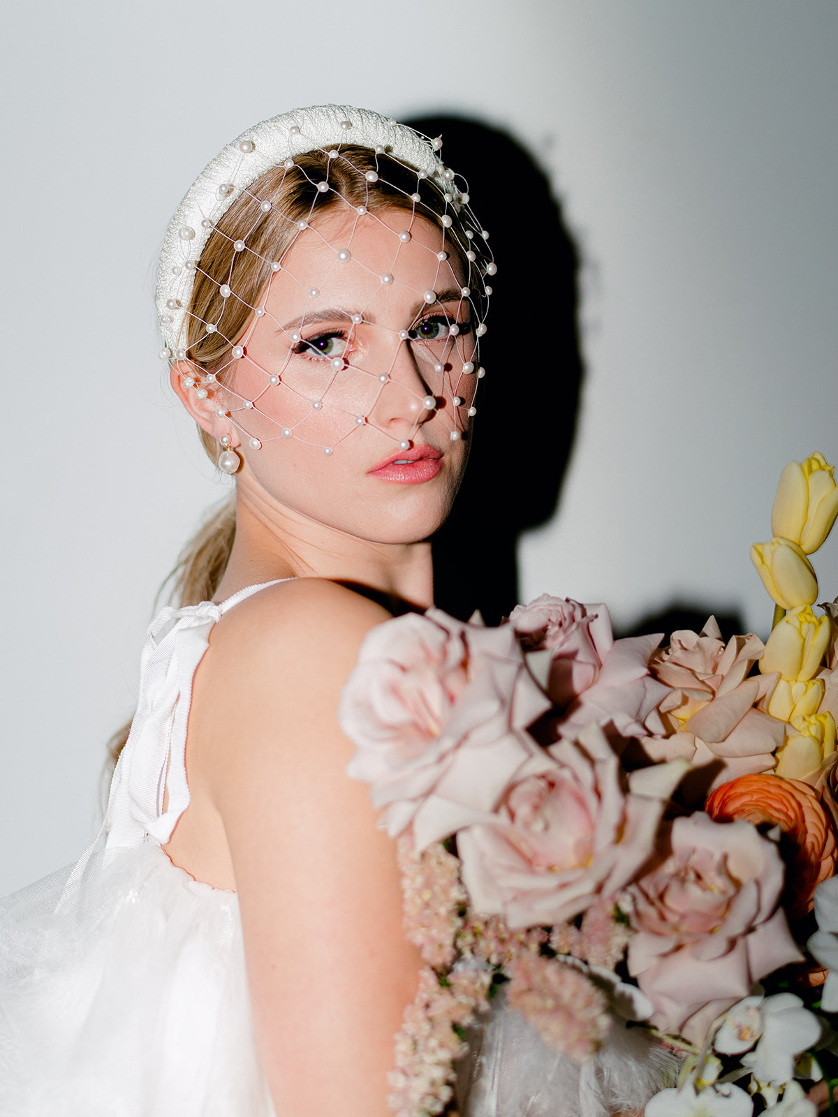Dynamic flash photography beautifully illuminating bright florals in captivating bridal portraits, a mix of modern and vintage aesthetic, portrait featuring a birdcage veil and white headband worn by a bride holding her bouquet in a feather wedding gown, low pony-tail bridal hairstyle