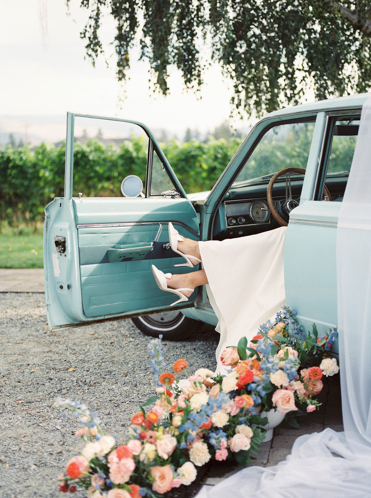 lush floral arrangements adorning a classic car, transforming it into a stunning centrepiece for a vintage-inspired wedding, bride wearing white satin bridal heels by Bella Belle Shoes kicks her feet up in a vintage baby blue car, unique bridal portrait inspiration