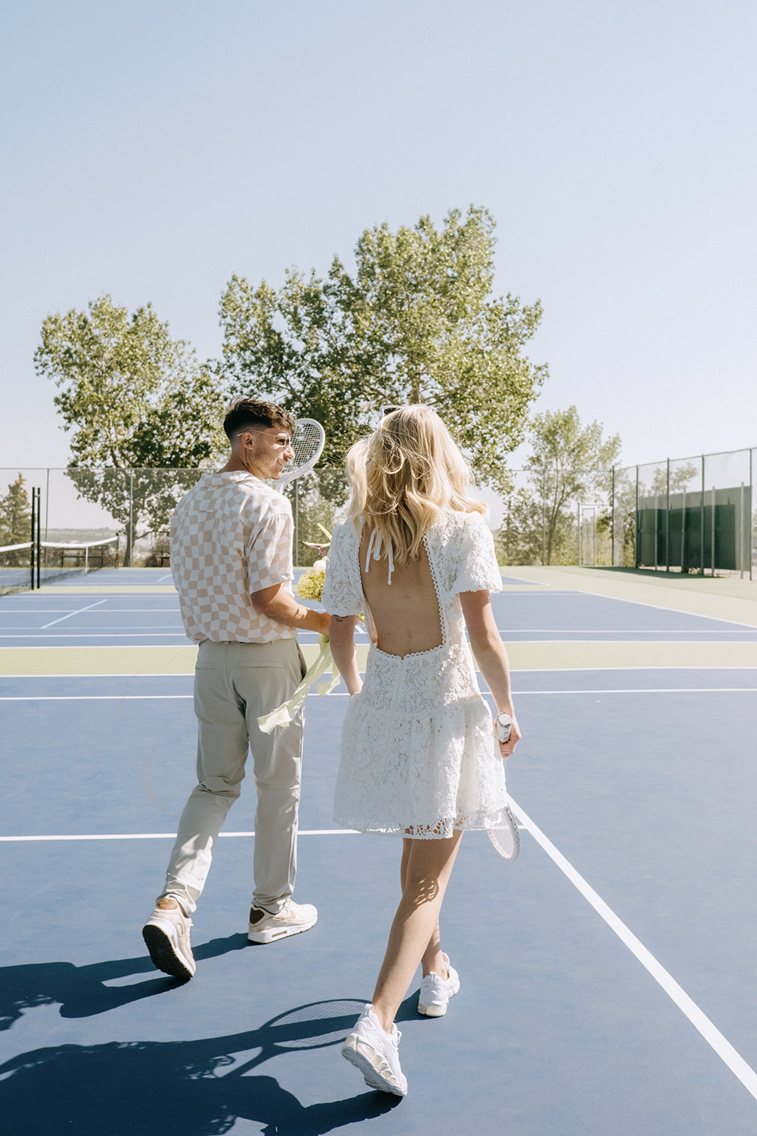 candid couple's portrait on tennis court during unique elopement, open-back short lace wedding dress by Asos with white bridal sneakers, casual wedding attire: alternative and sporty wedding ideas