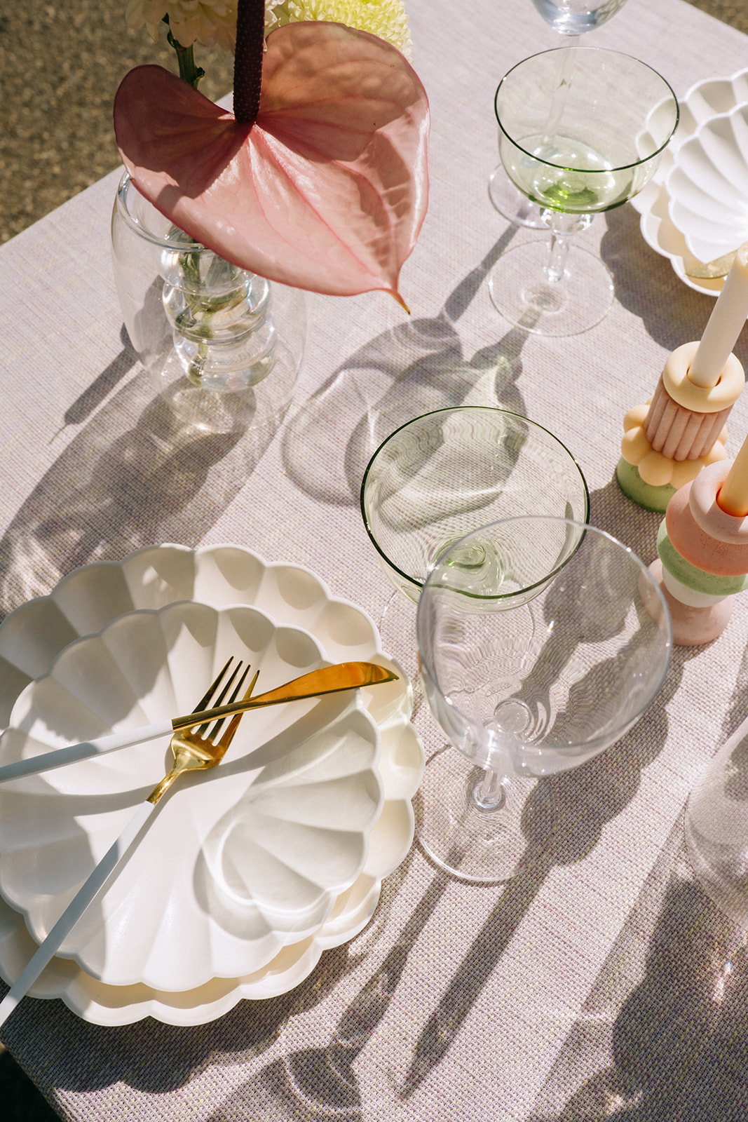 Pink linen tablecloth featuring chartreuse coup glasses and gold cutlery, unique white scalloped plates for outdoor wedding reception, pastel candlesticks in summer-inspired colour palette of pastel greens, pinks and oranges