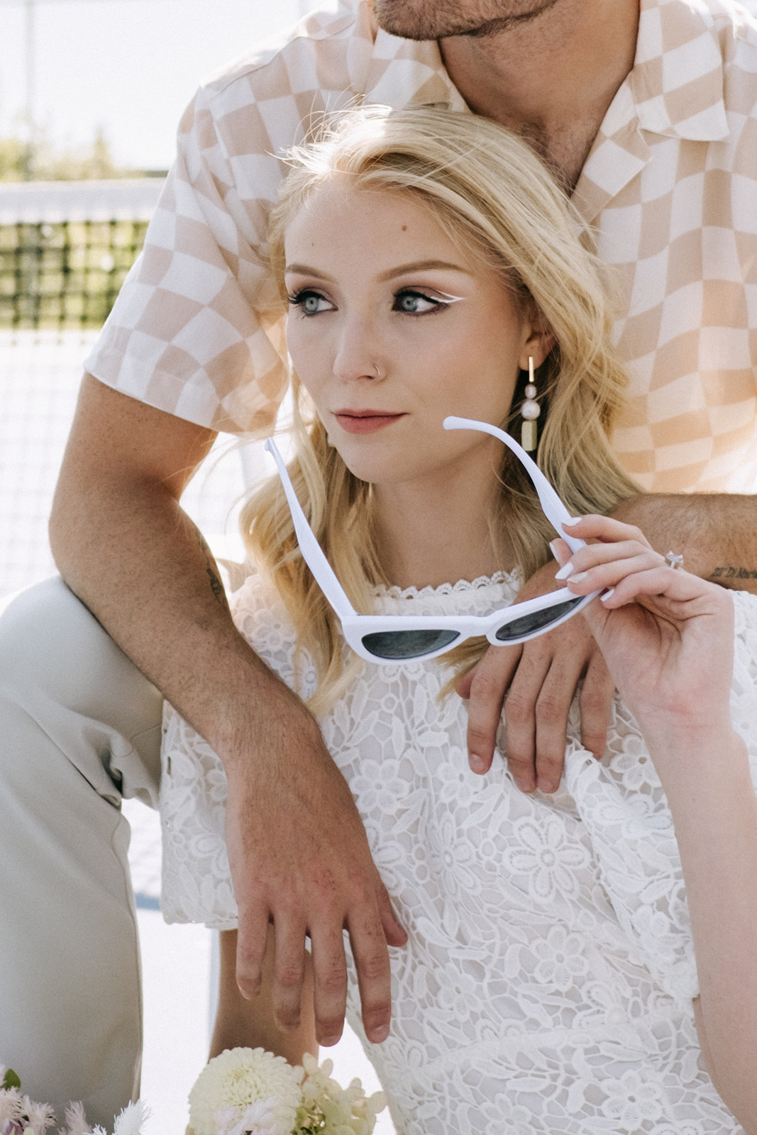 Bridal portrait on tennis court with bride wearing white cat-eye sunglasses, alternative white eyeliner bridal make-up inspiration, casual groom attire for couples portraits on a tennis court