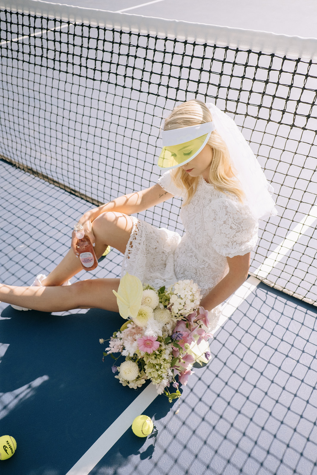 Short bridal veil and clear chartreuse tennis visor for a fashion -forward bride, bridal portrait leaning against tennis court net showcasing bouquet by Hen and Chicks in neons and pastel colour palette, short wedding dress ideas for casual elopement