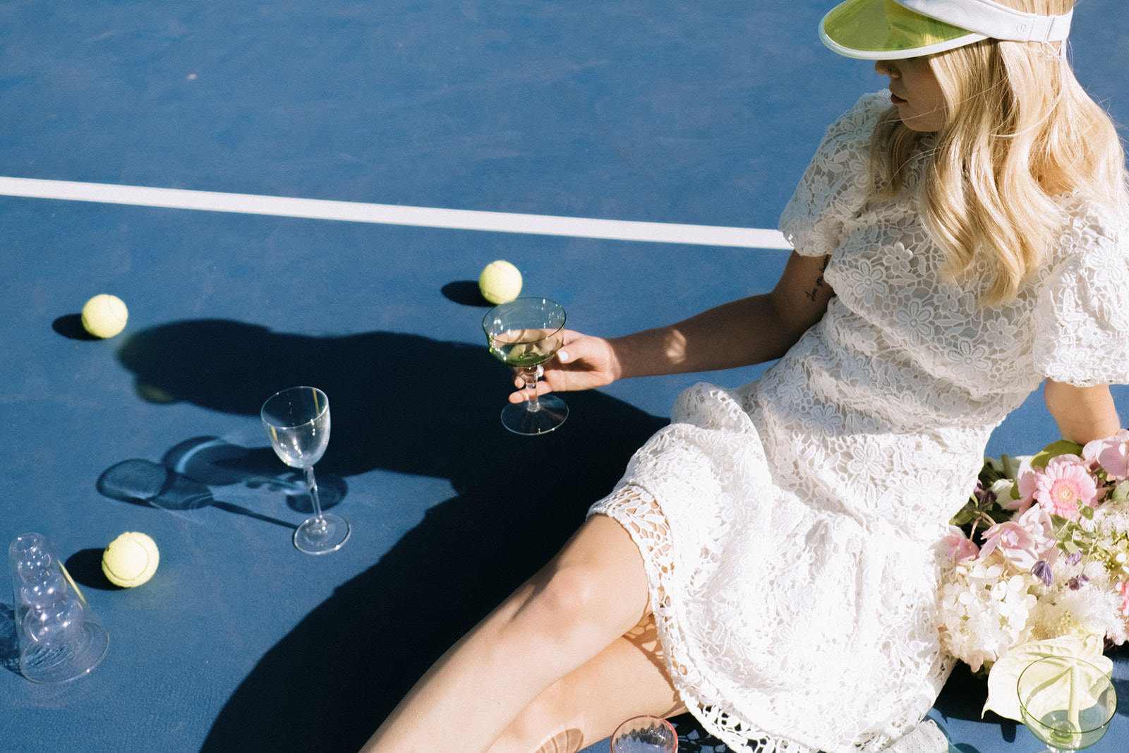 Bride enjoying a casual and laid-back cocktail during elopement, fashion-forward bridal style featuring short white lace wedding dress and tennis visor, pastel pink, white and chartreuse bridal bouquet