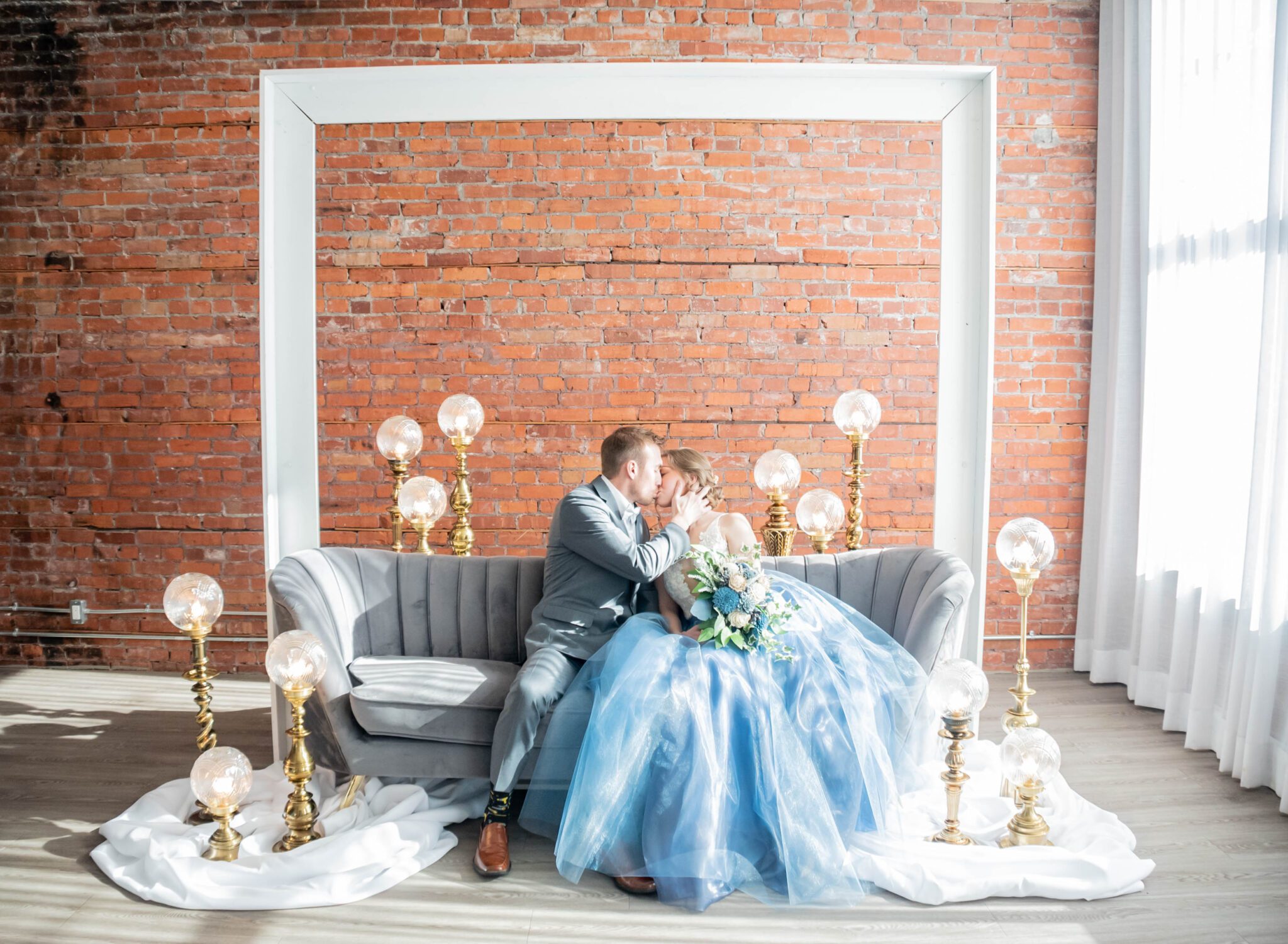 Vintage lighting surrounds bride and groom sharing intimate moment on a grey velvet couch in a unique Calgary, Alberta wedding venue, groom is wearing a timeless and traditional grey suit, bride is holding large bouquet full of blue, peach and white blooms