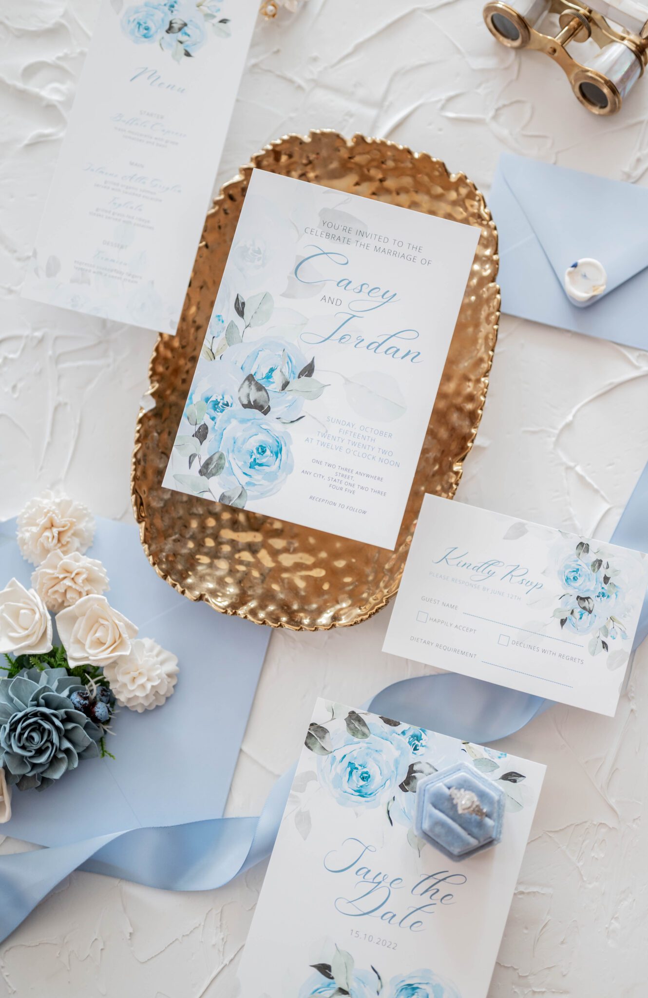 Flat-lay photo featuring french-inspired custom invitations, save-the-dates, RSVP card and menu by Red Fox Multimedia, white and blue floral accents on top of baby blue envelopes with white wax seal, romantic stationery ideas in blue