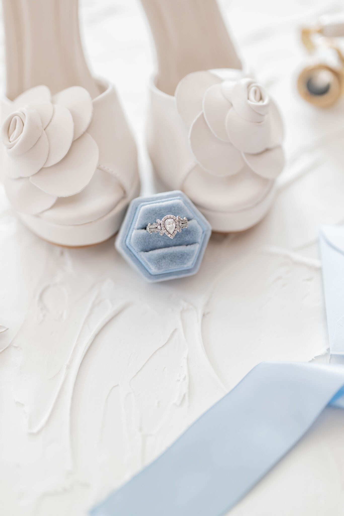 Flat lay photo of baby blue velvet ring box with pear shaped engagement ring, white leather bridal heels with large white flower on the toe, baby blue ribbon detail on a white plaster table top