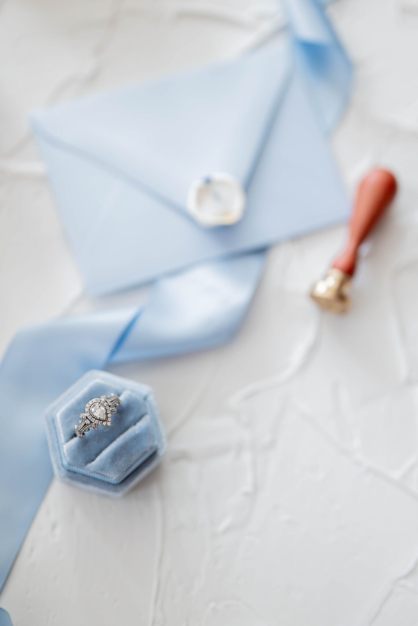 Flat lay photo featuring vintage pear engagement ring in baby blue velvet box, blurred background showing baby blue ribbon accents, baby blue envelope with a white wax seal and brass stamp, plaster table top design ideas