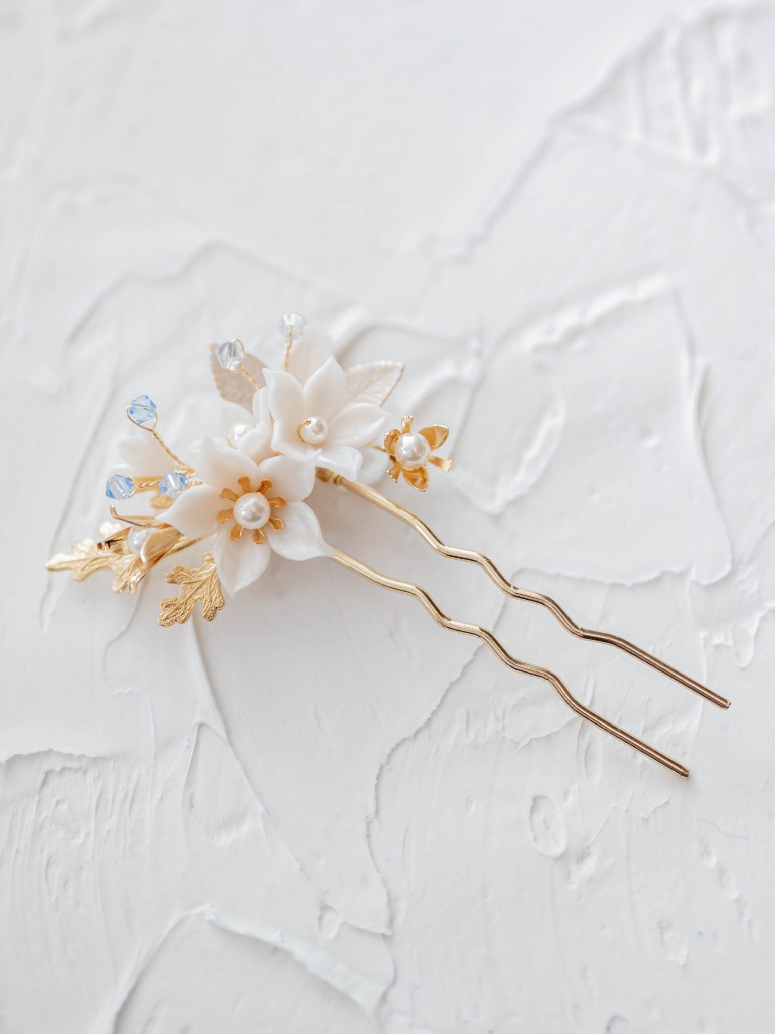 detailed photo of gold hair pin featuring white flowers, gold leaves and small baby blue accents, flat lay photography using plaster background, something blue bridal hair accessory inspiration