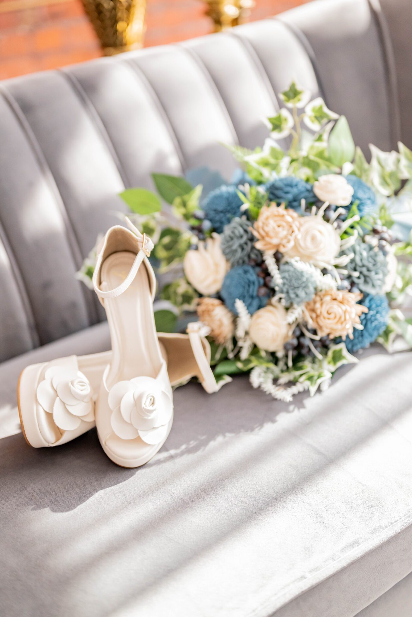 Vintage white bridal heels with ankle strap and large flower on the toe next to baby blue and peach bouquet with ivy accents, detail photo of bridal shoes and bouquet on grey velvet couch