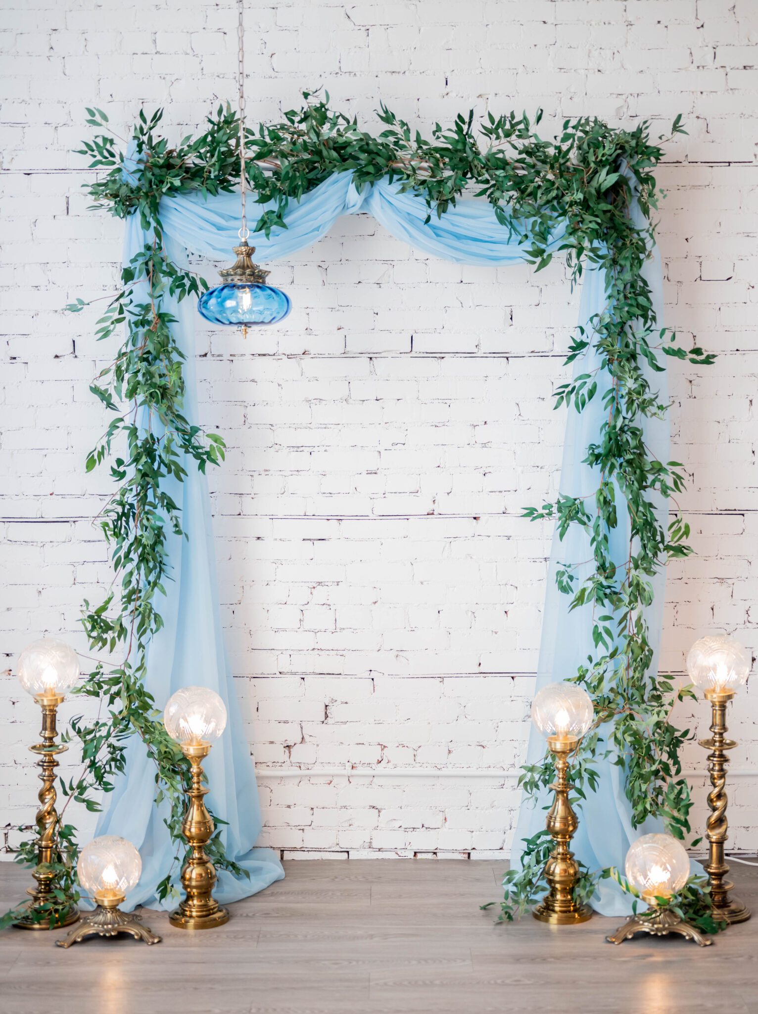 Vintage lighting decor ideas for a romantic ceremony, greenery and baby blue draping on ceremony arch in front of white brick wall, french-inspired with a modern twist wedding inspiration in the heart of Calgary, Alberta