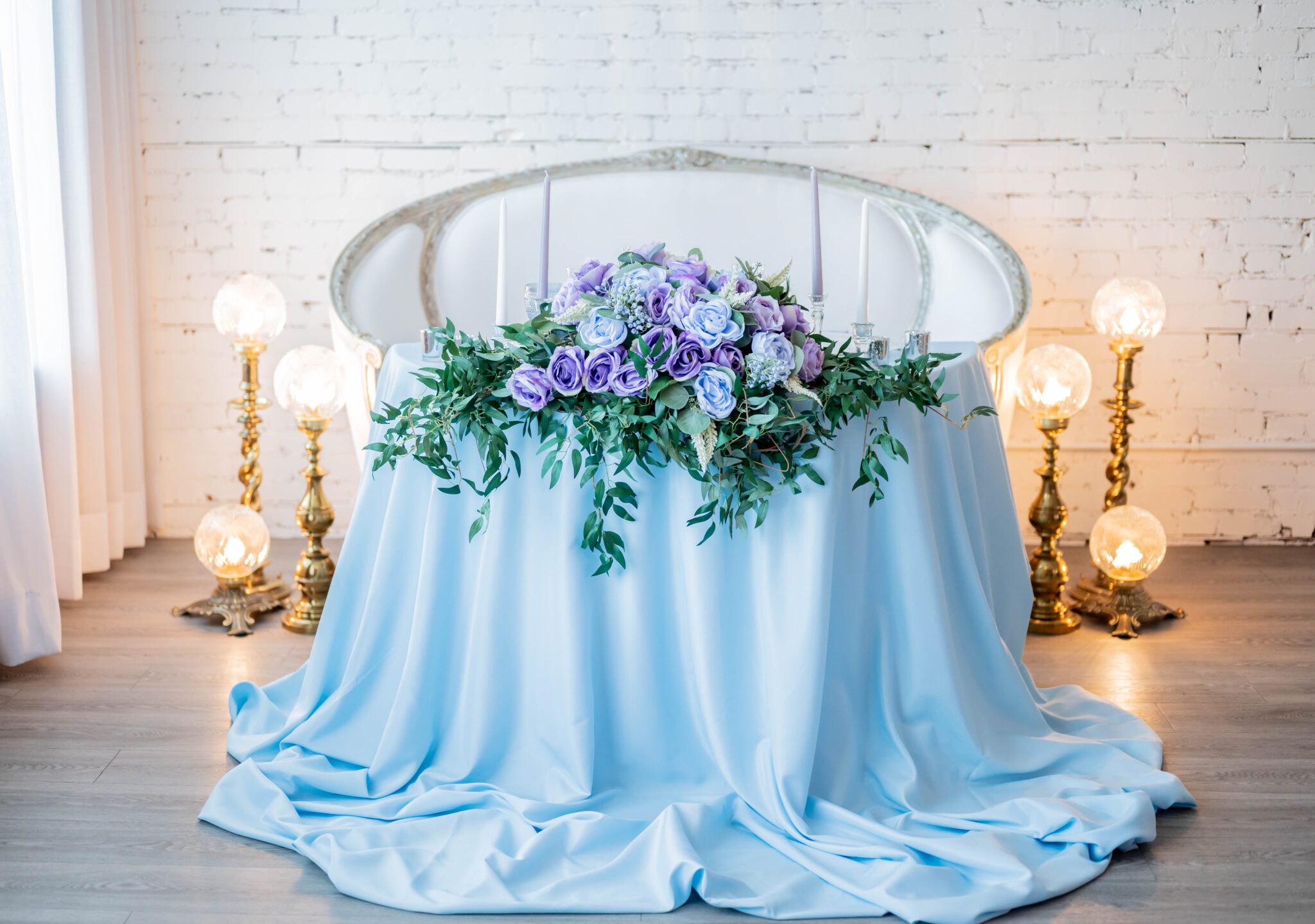 Elegant sweetheart table with lush baby blue and purple florals and greenery, vintage brass lighting for a romantic ambiance during reception, white and silver love seat for bride and groom