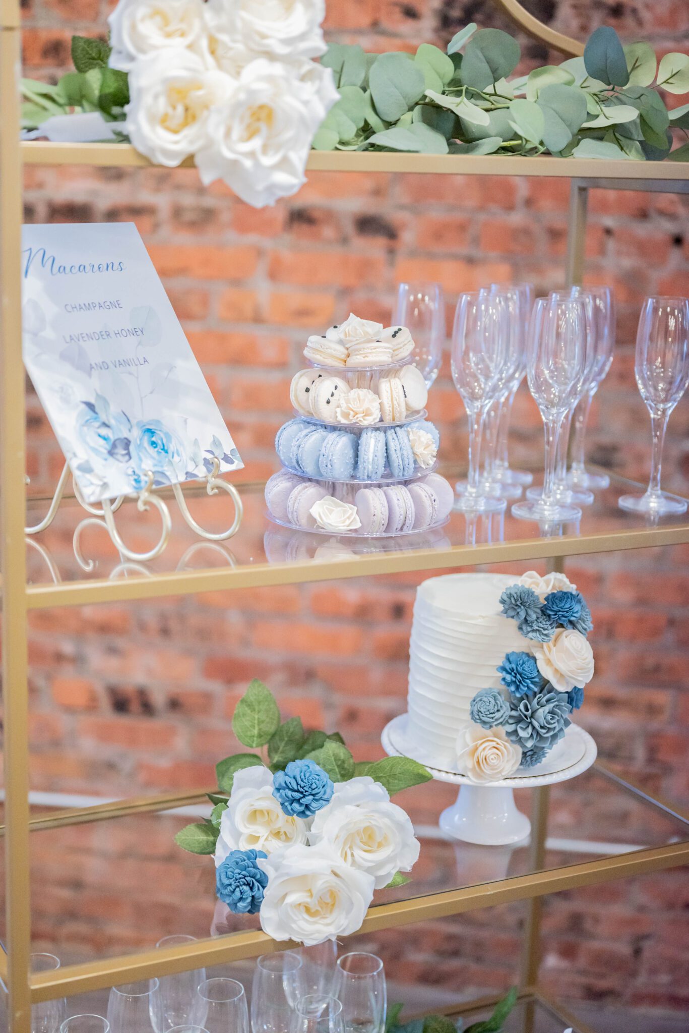 tiered dessert stand displaying light purple, baby blue and creamy white french macarons, white wedding cake decorated with white and blue florals, custom dessert sign with blue roses and grey-toned on vintage white easel, gold frame and glass shelf dessert and champagne display ideas