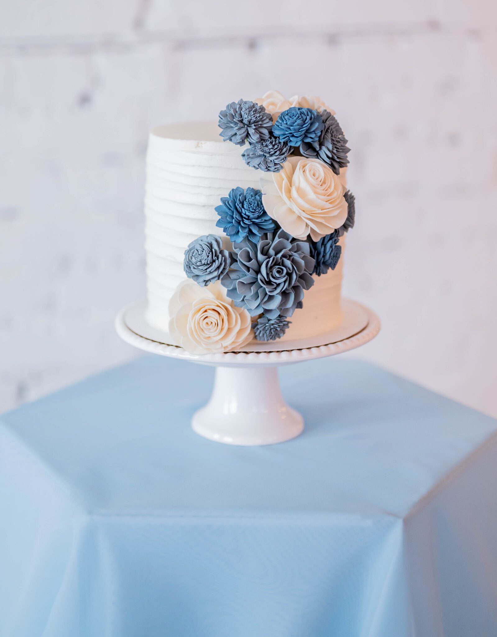 Simple and classic cake table with white cake stand and baby blue linen, single-tiered white cake with white, baby blue and dark blue edible flower decorations by Cakes by Cata