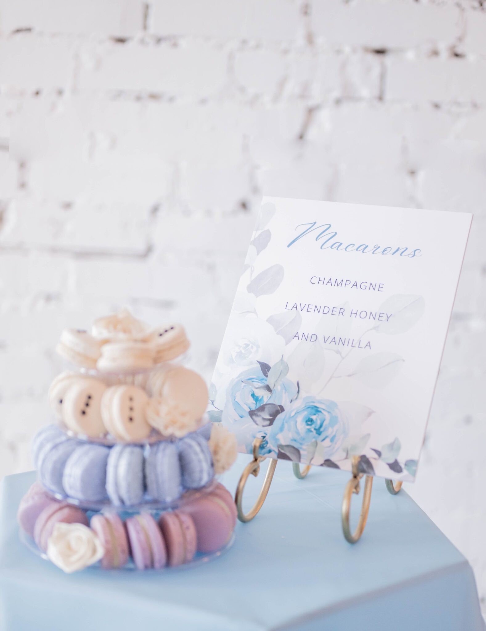 French macaron dessert ideas for wedding guests by Ollia Macarons & Tea, tiered display of macarons in purple, baby blue and creamy white decorated with white florals, dessert table with blue linen and custom dessert sign sitting in a vintage brass easel