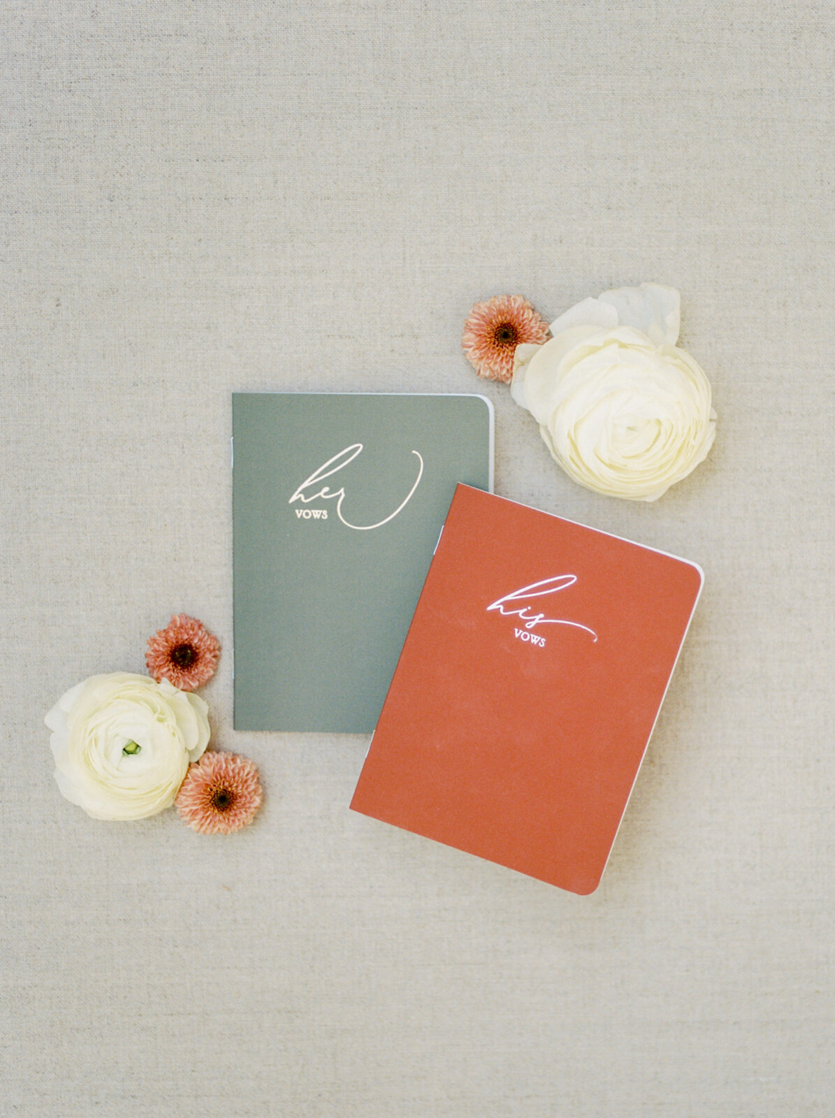 His and hers vow books in muted shades of orange and green for a fall wedding colour scheme, white florals with accents of orange, modern flat lay design with vow books and florals