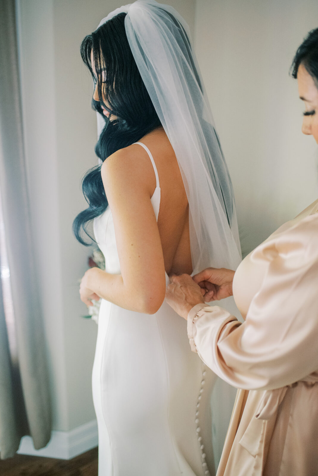 low-back bridal gown with thin straps and small buttons down the back, getting ready photo featuring mother-of-the-bride fastening wedding gown