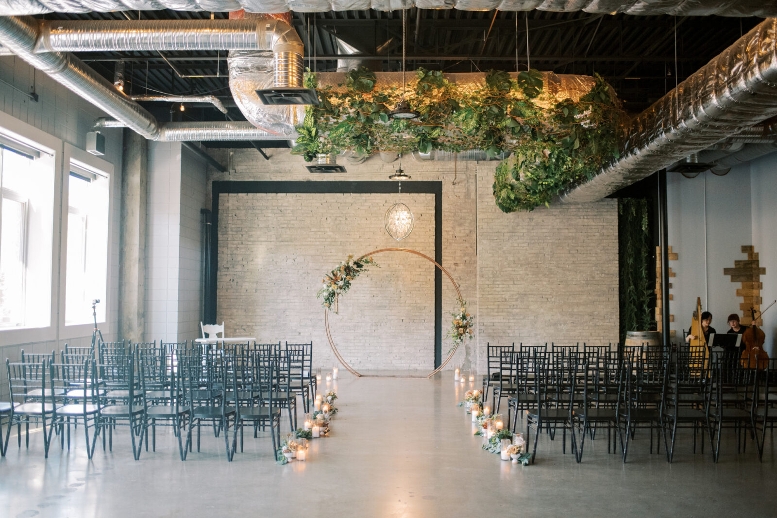 Industrial wedding ceremony at The Foundry Room in Edmonton, Alberta, gold circular arch with fall-toned florals and greenery at the end of the aisle, black chiavari guest chairs for modern and classic ceremony style