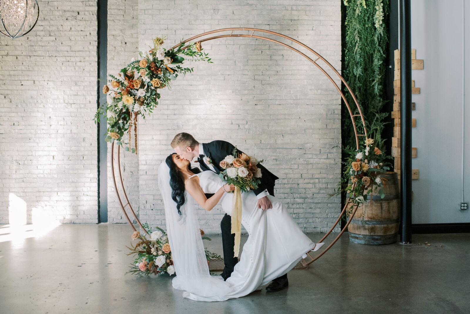 Modern and contemporary gold circular arch for wedding ceremony, florals in fall colour palette for wedding arch, bride and groom first kiss at indoor ceremony
