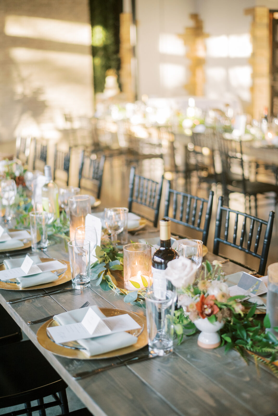 black chiavari chairs for guests during wedding modern wedding reception, fall wedding florals with lush greenery on guest tables, gold charger place settings on long wood tables rented from Tarnished Timber