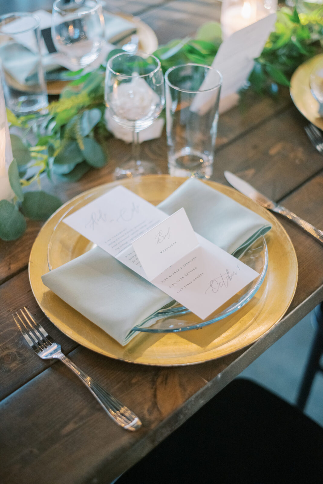 gold charger place settings on long wood tables for fall wedding, custom wedding menu and place card stationery for modern and contemporary wedding, lush greenery centrepieces for fall wedding