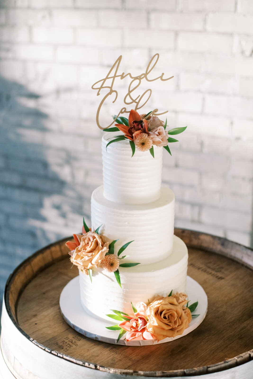 Tiered white wedding cake featuring florals in a fall themed palette by The Art of Cake, three tiered wedding cake inspiration, delicate custom cake topper of the bride and groom's names