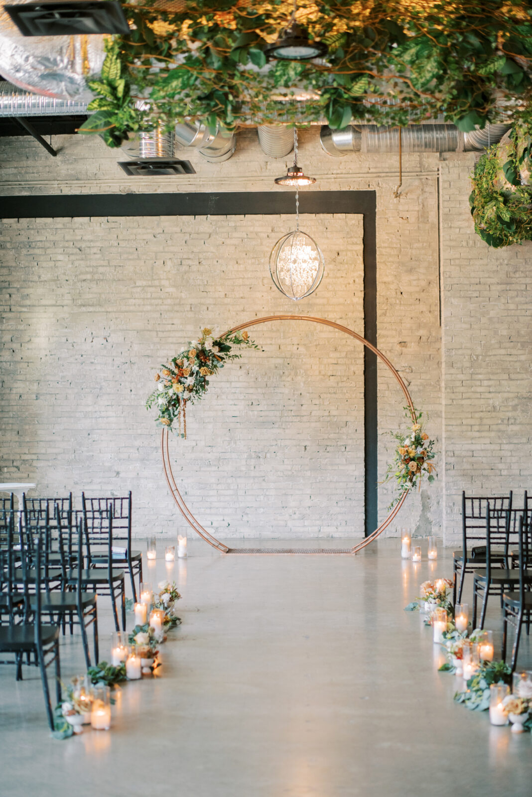 black chiavari guest chairs for modern and classic ceremony style, gold circular arch with fall-toned florals and greenery at the end of the aisle, hanging greenery installations for indoor wedding ceremony