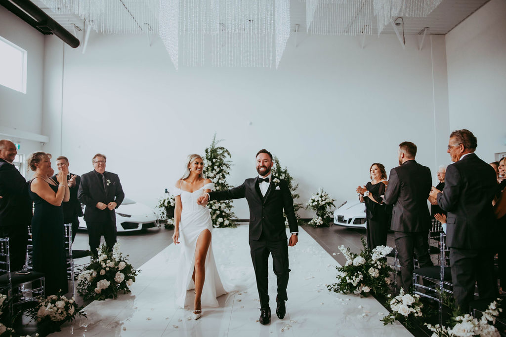 Modern, glam Red Deer wedding at unique car dealership venue. Bride and groom exiting wedding ceremony, surrounded by family and friends in black wedding attire, black and white wedding inspiration. 