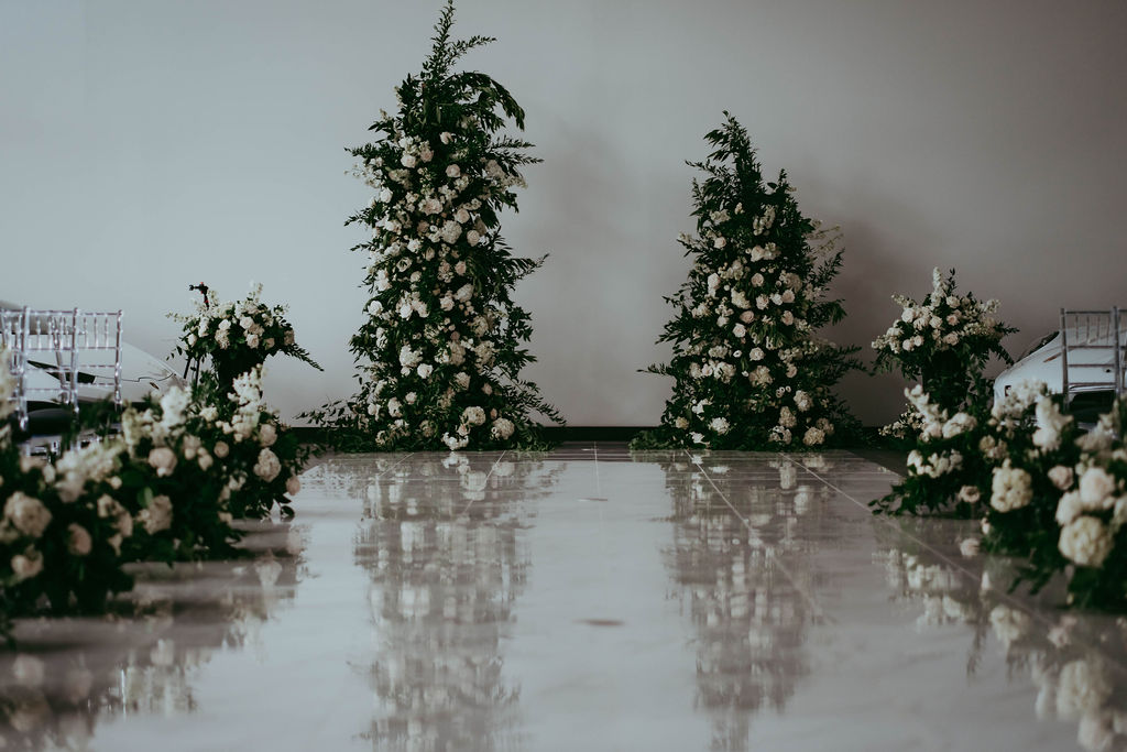 A row of elegant floral arrangements with white florals and greenery, leading to primary floral arch in Red Deer wedding ceremony space. 