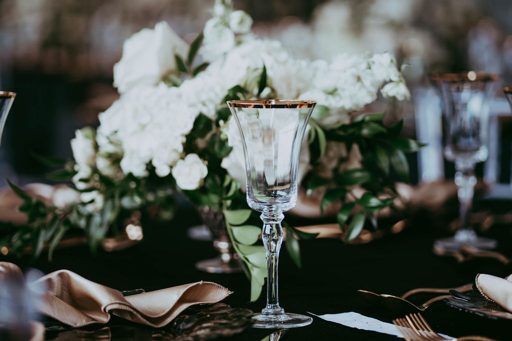 Elegant gold-rimmed clear glassware with silk gold napkins, gold coloured flatware on modern black linen, centrepiece vase with white florals and added greenery. 