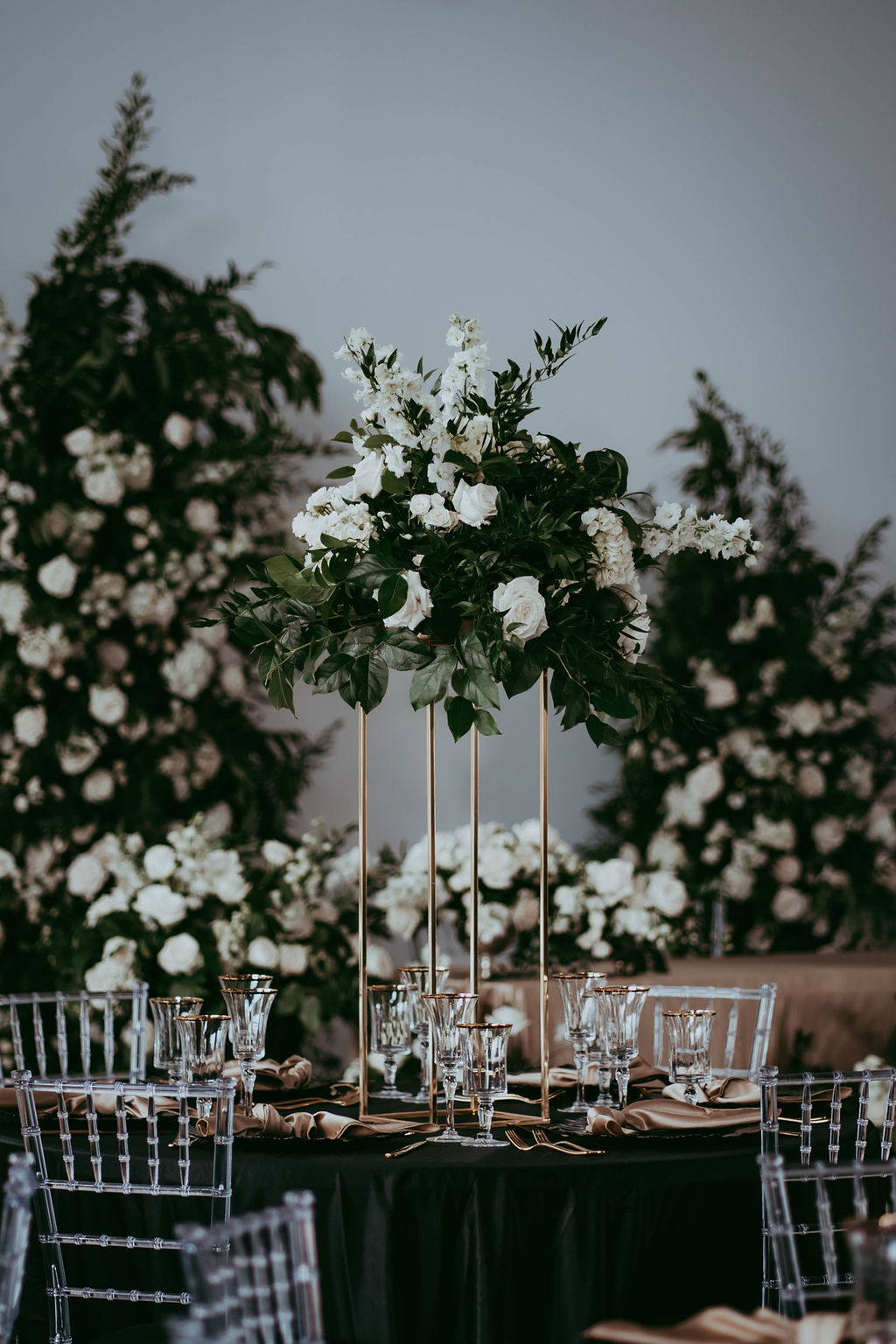 Black table linens, clear chiavari chairs, silk gold napkins, gold-rimmed clear glassware, modern arrangements with white florals and greenery, black and white wedding inspiration. 