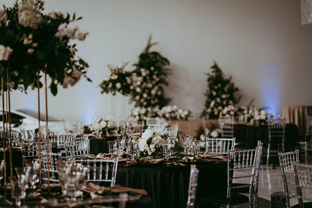 Black and white reception details with modern tablescape, large floral arrangements on tables with clear chiavari chairs, gold-rimmed glassware, and black tablecloths. 