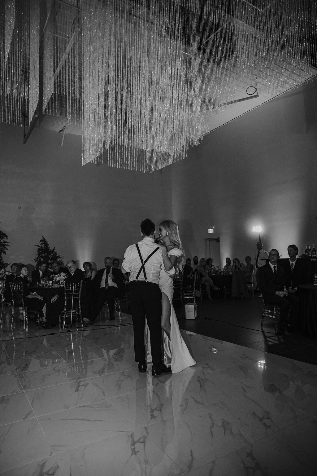 Bride and groom sharing their first dance on a glossy, marble dance floor in front of seated guests at their Red Deer wedding, glam wedding inspiration, bride wearing modern gown with slit. 