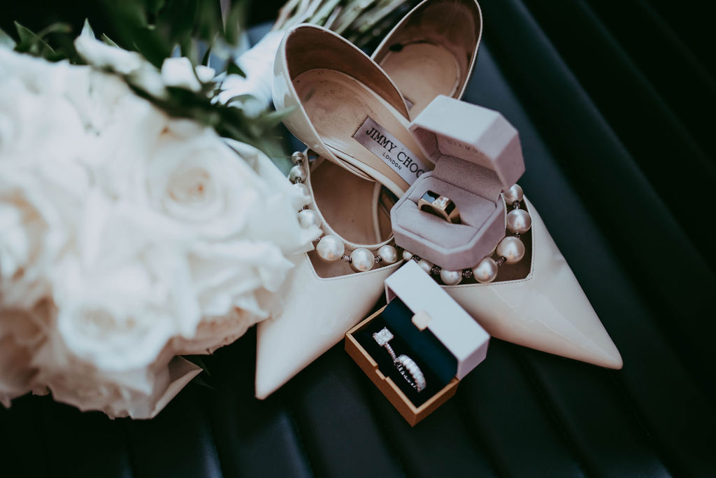 Elegant getting ready flatlay with pearl detailing on Jimmy Choo bridal shoes, surrounded by white florals and couple's wedding rings. 