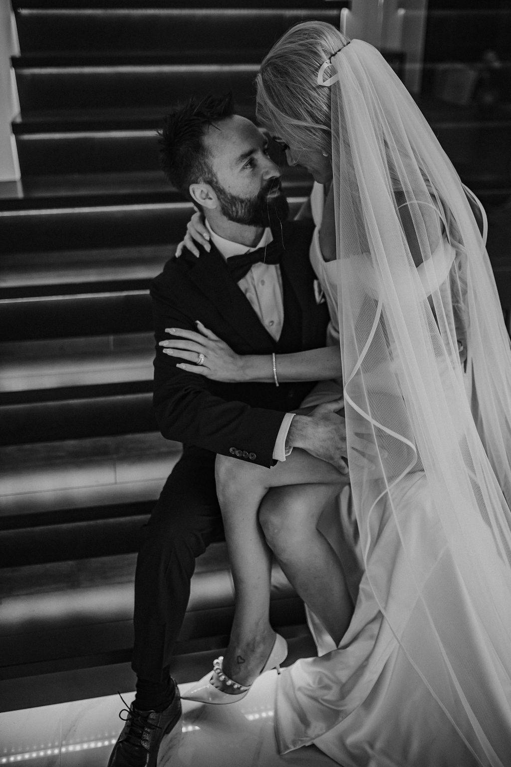 Groom holding bride while her wedding gown with a slit drapes to the side, sitting on modern staircase in Red Deer wedding venue. 