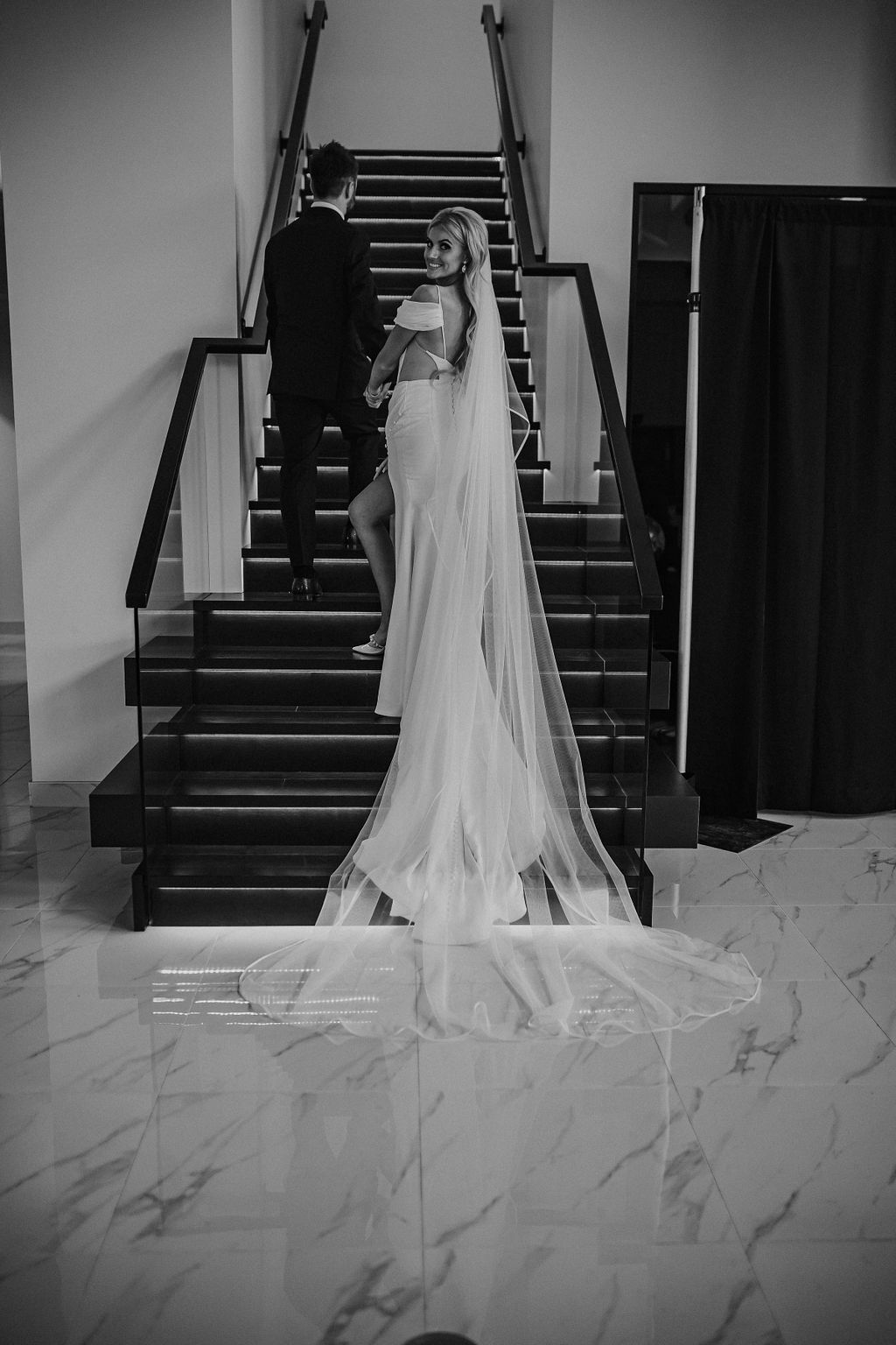 Modern, glam Red Deer wedding at unique car dealership venue. Portrait of groom leading his bride up the black, elegant staircase. Bride looking back while her wedding gown with slit drapes down the stairs. 