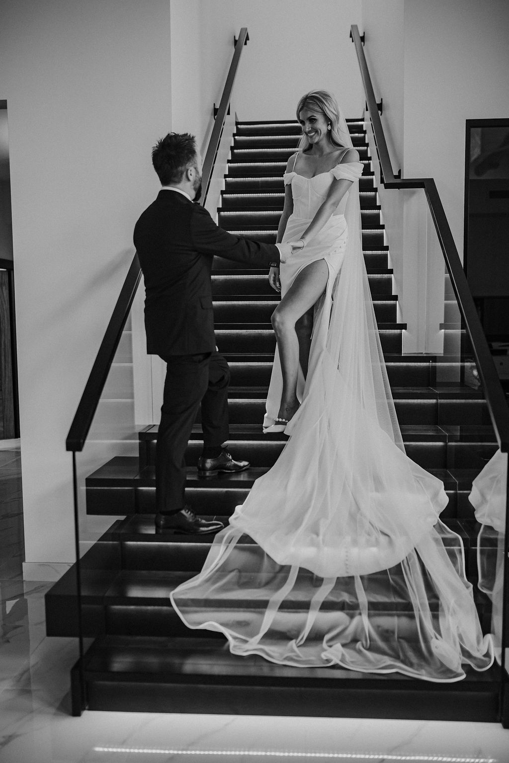 Groom leads bride down the dramatic staircase in modern, Red Deer wedding venue, bride is wearing an elegant wedding gown with slit providing a contrast to the black staircase. 