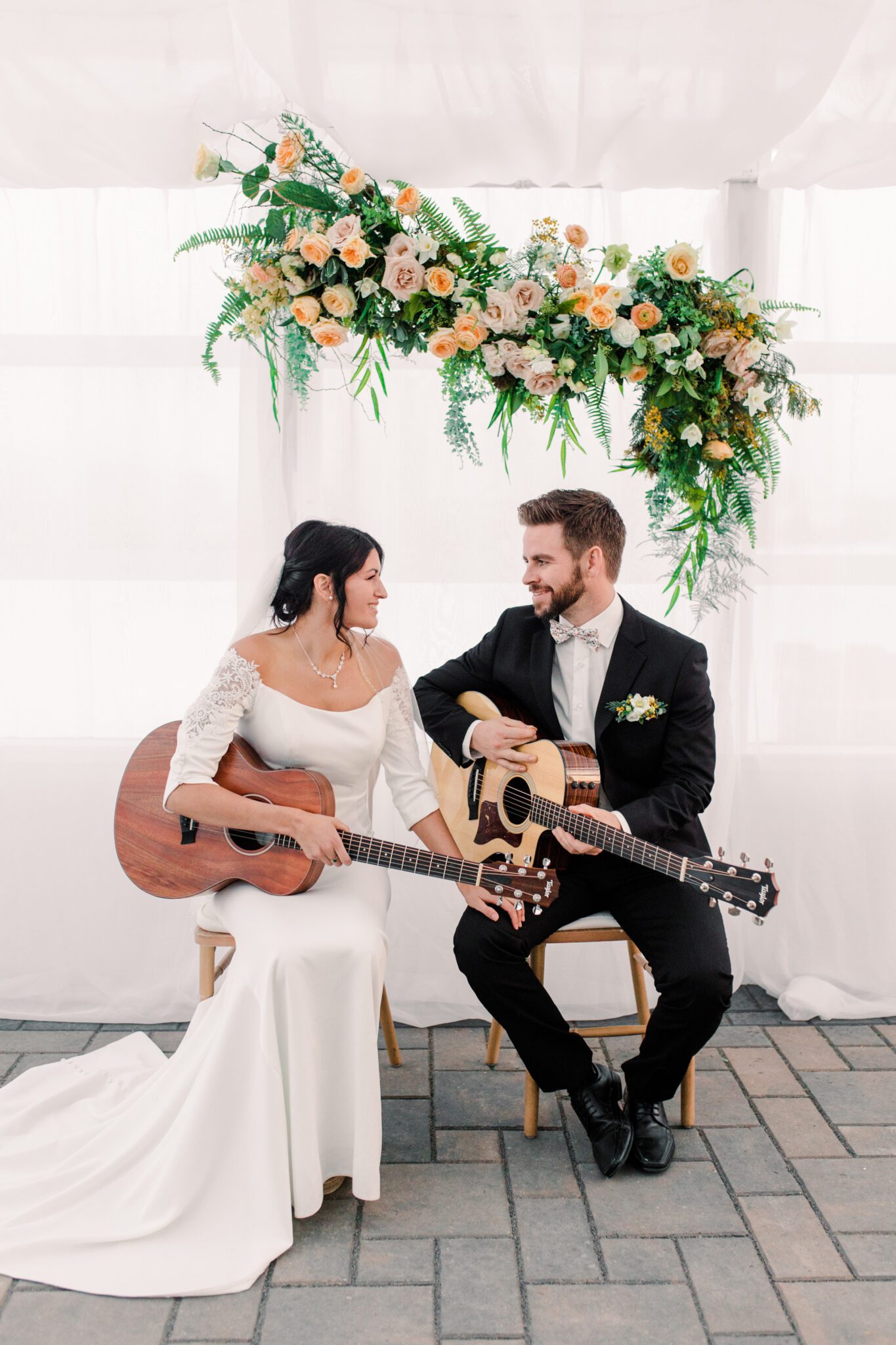 Fun bride and groom playing guitar at summer tented wedding reception, spring inspired floral installation designed by Little Petal Company featuring roses and cascading ferns