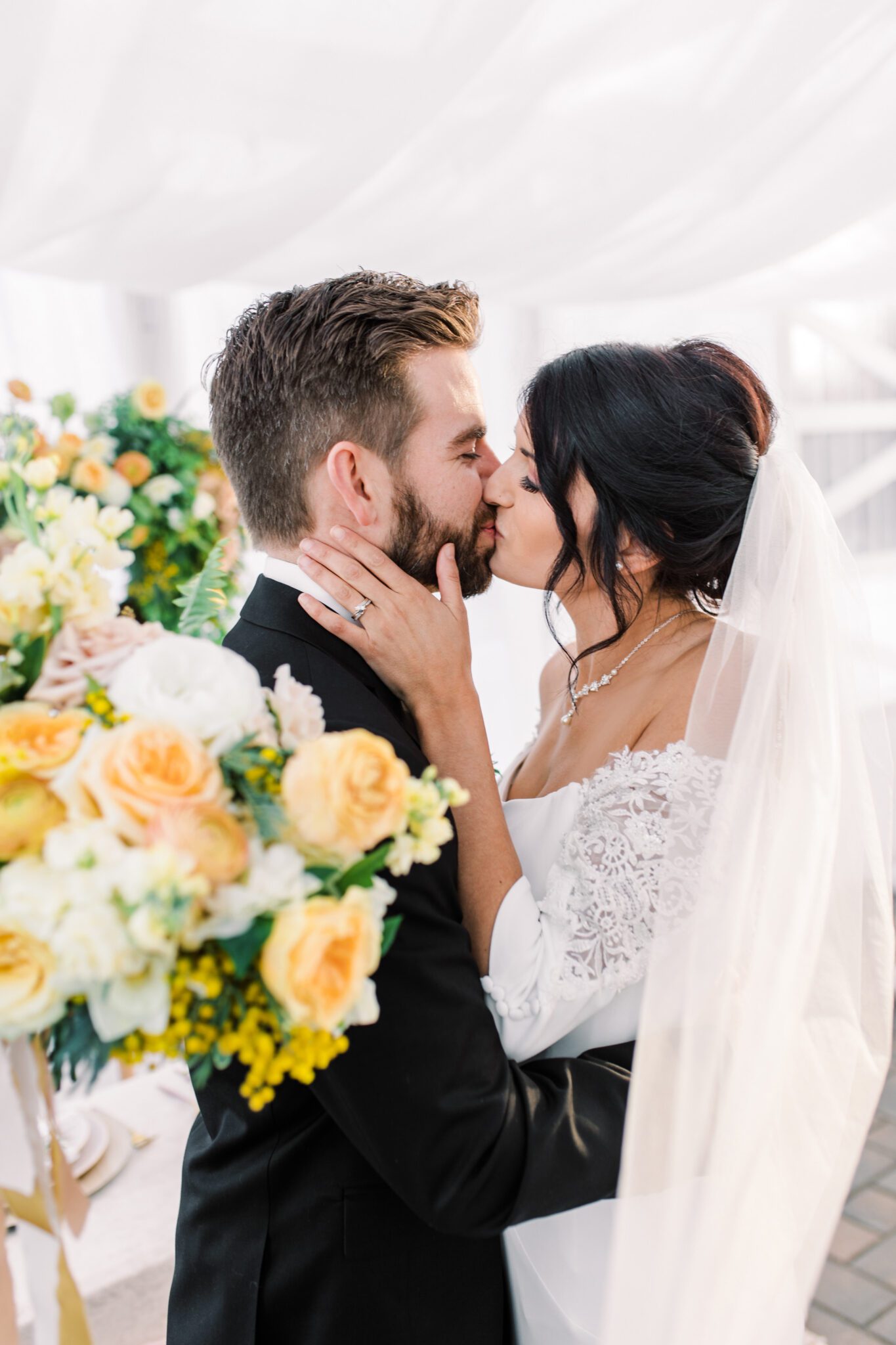 Bride and groom kissing at stunning tented wedding, featuring yellow and peach spring wedding colour palette. Bride wearing classic off-the-shoulder wedding gown with lace sleeve detail.