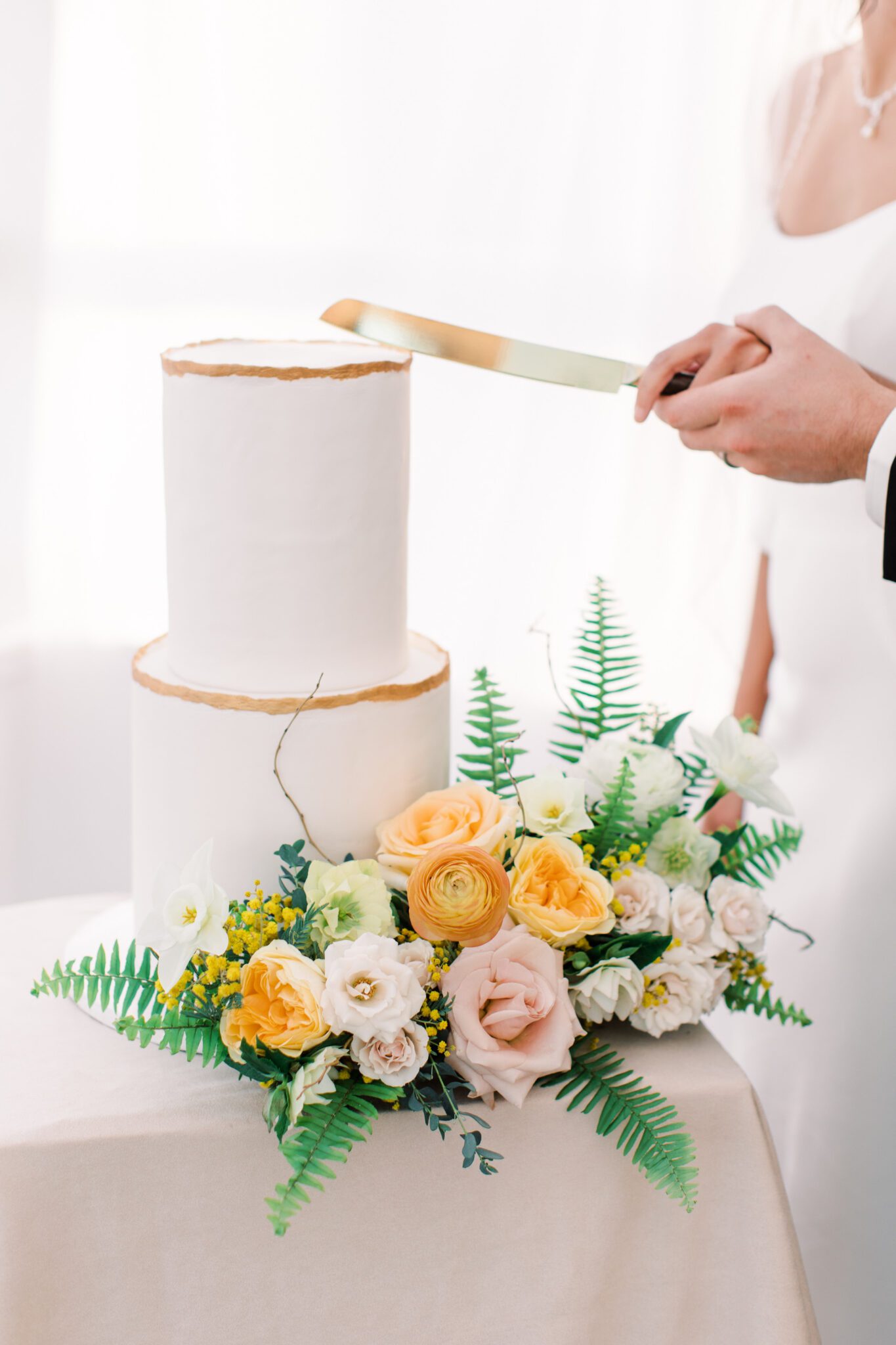 Couple cutting classic and elegant two-tiered white wedding cake with gold detail by Pink Oven