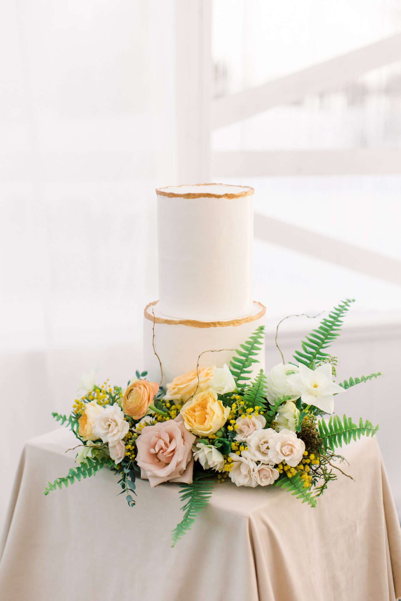 Classic and elegant two-tiered white wedding cake with gold detail by Pink Oven, surrounded by yellow and peach spring florals.
