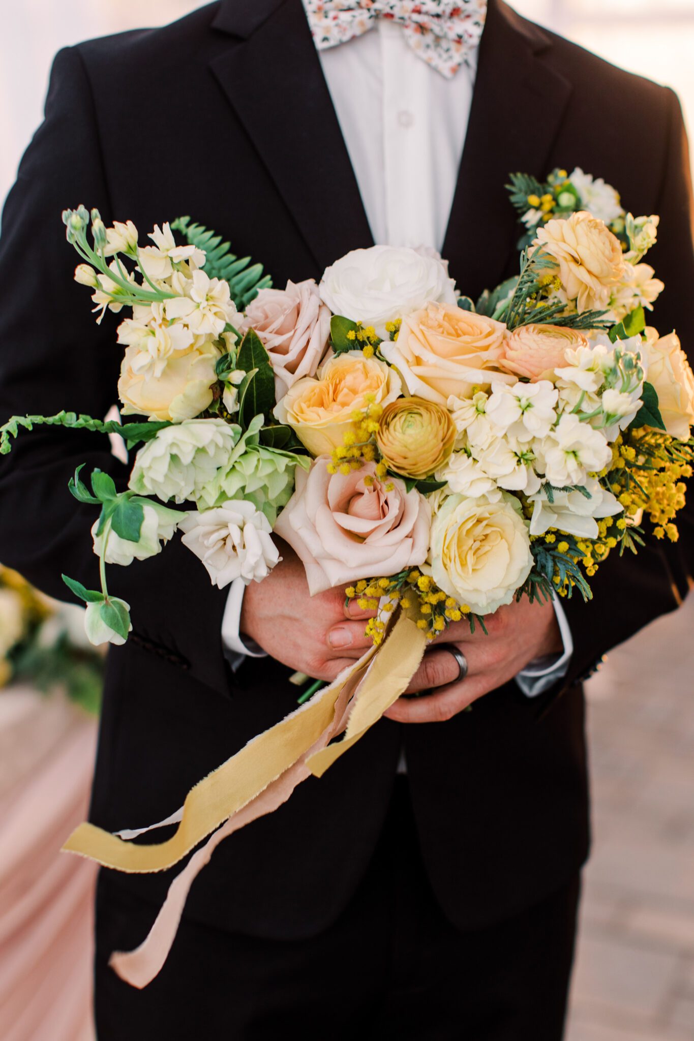 Groom holding stunning yellow and pink spring inspired wedding bouquet, wearing floral bowtie from Edward's Factory Outlet
