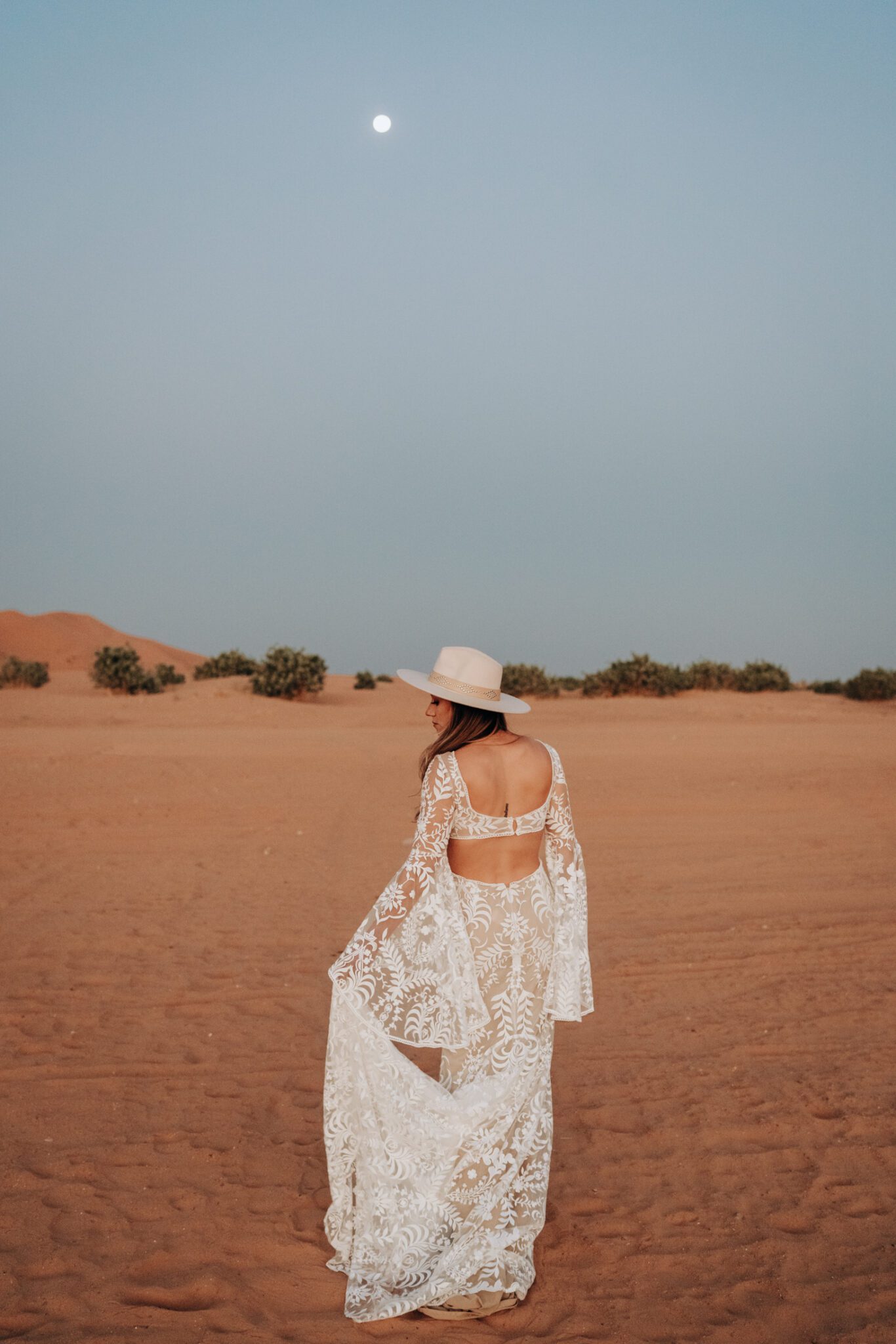 Bohemian bride wearing lace gown standing in the sand during her moroccan desert engagement session.