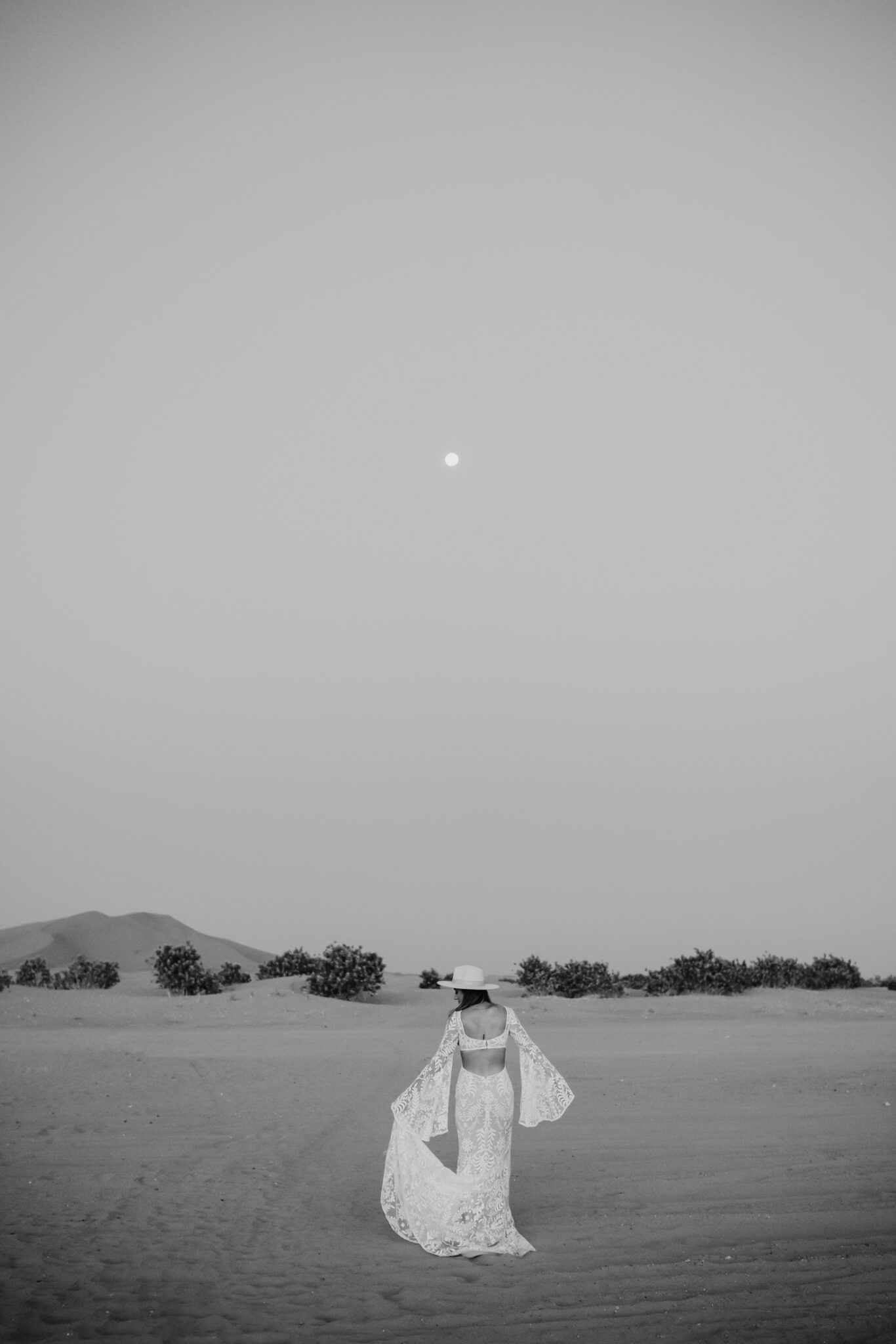 Bohemian bride wearing lace gown standing in the sand during her moroccan desert engagement session.